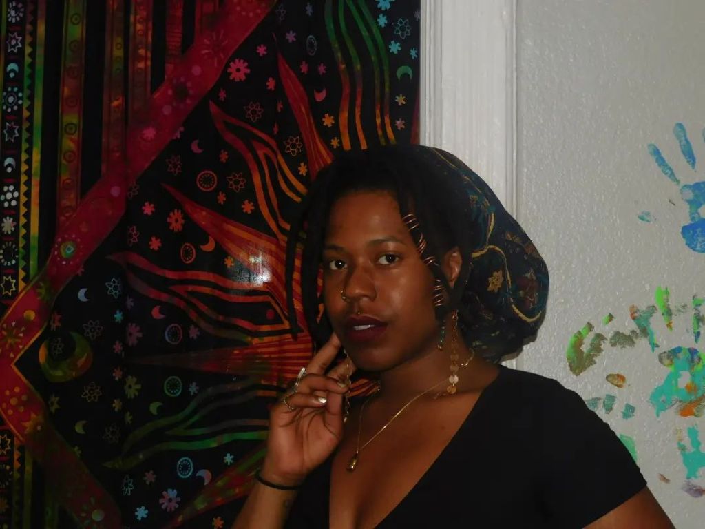 A woman stands before a vibrant tapestry filled with sun, moon, and flame motifs. Her head is wrapped in a patterned scarf, and her expression is serene yet intense. One hand gently touches her lips, adorned in gold jewelry and dark lipstick, as if caught mid-thought or spell.