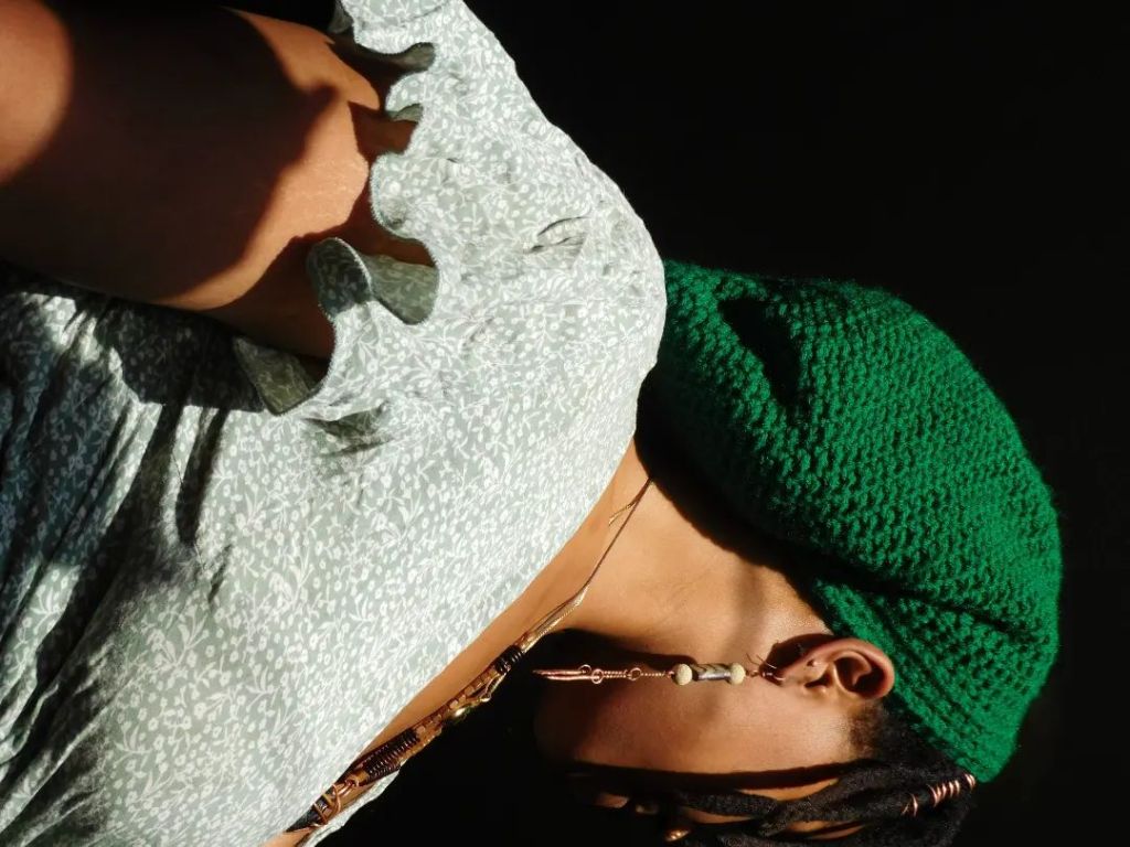 A woman is turned away in profile, head tilted downward in a moment of stillness. She wears a soft green ruffled dress and a crocheted emerald headwrap. Sunlight brushes across her skin, highlighting her collarbone and the curve of her arm, casting deep shadows behind her.