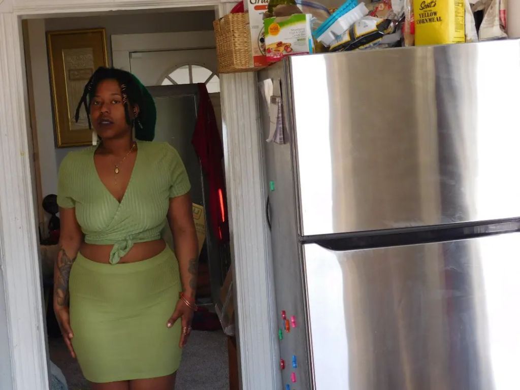 A woman stands in a kitchen doorway wearing a sage green wrap top and skirt. Sunlight filters in, casting soft shadows. A stainless steel refrigerator occupies the foreground, cluttered with food items on top.