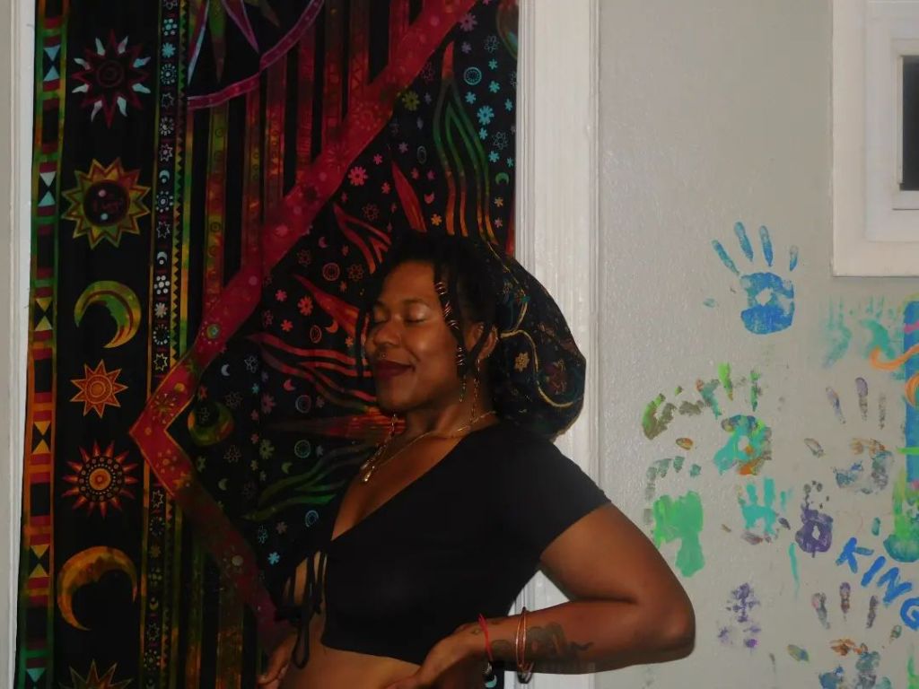 A woman in a black crop top stands with her eyes closed and a soft smile, framed by a colorful cosmic tapestry and a wall dotted with childlike handprints. Her hair is adorned with a beaded scarf wrap, and her posture is confident, grounded, and serene.