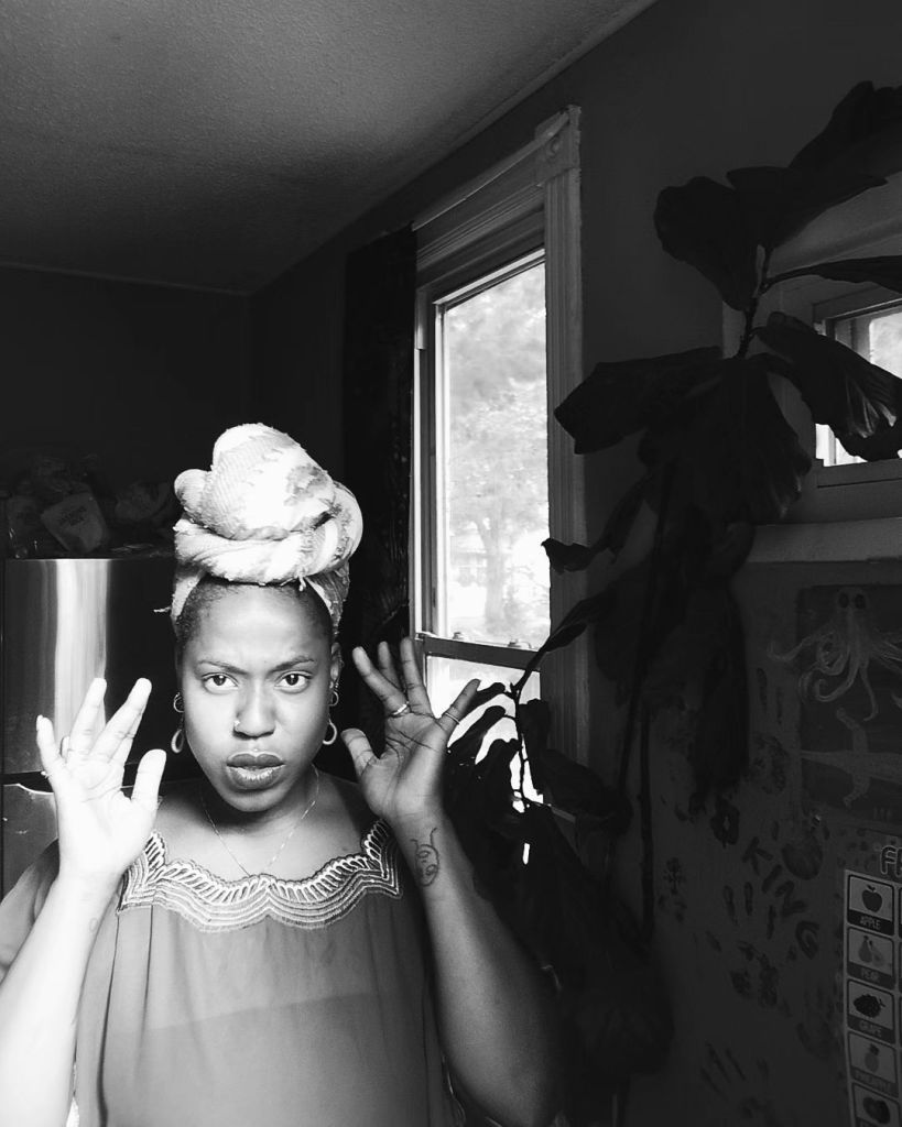 A black woman in a headwrap stands indoors near a window, hands raised mid-motion. The photo is in black and white, with soft light falling through the window beside her.