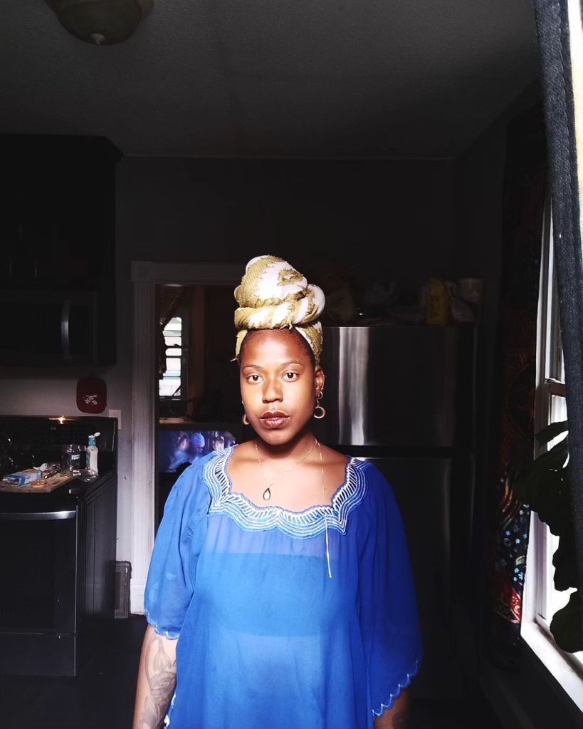 A woman stands centered in a kitchen, wearing a sheer blue garment and a golden headwrap. Sunlight spills in through a window beside her, casting soft highlights on her face and attire. She gazes forward, grounded and luminous.