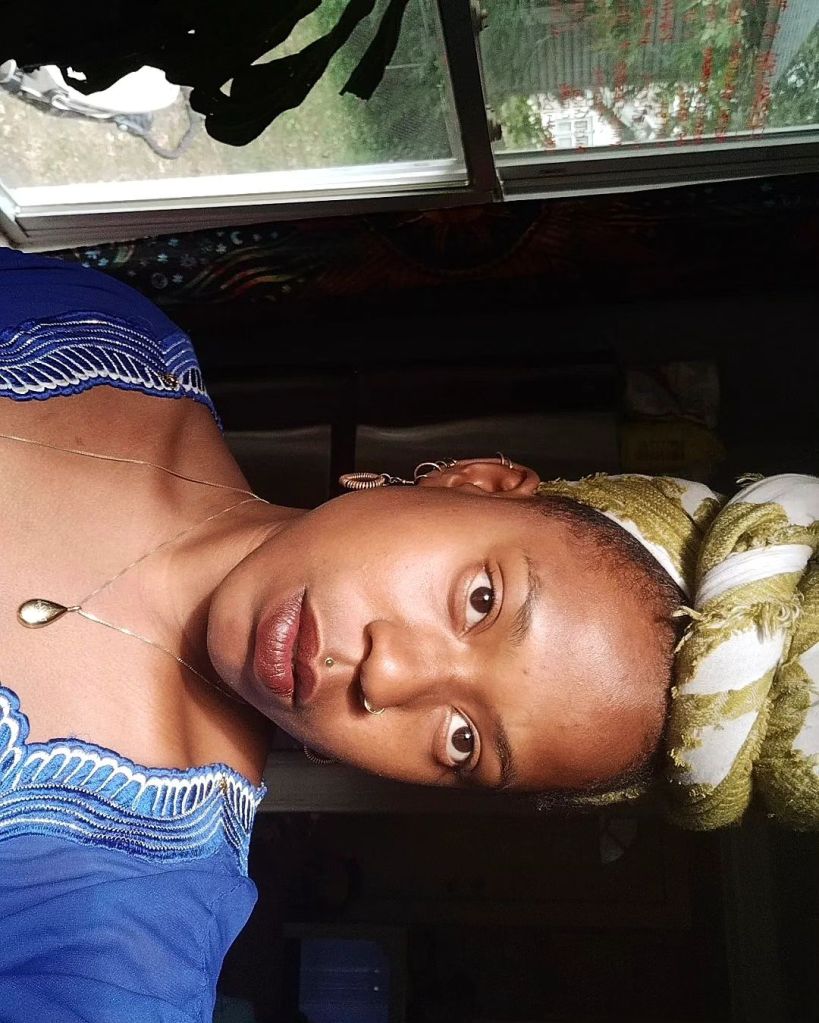 A woman in a royal blue dress and golden headwrap gazes directly at the camera, lit softly by natural window light. She wears a teardrop necklace and gold earrings, with greenery faintly visible behind the window.