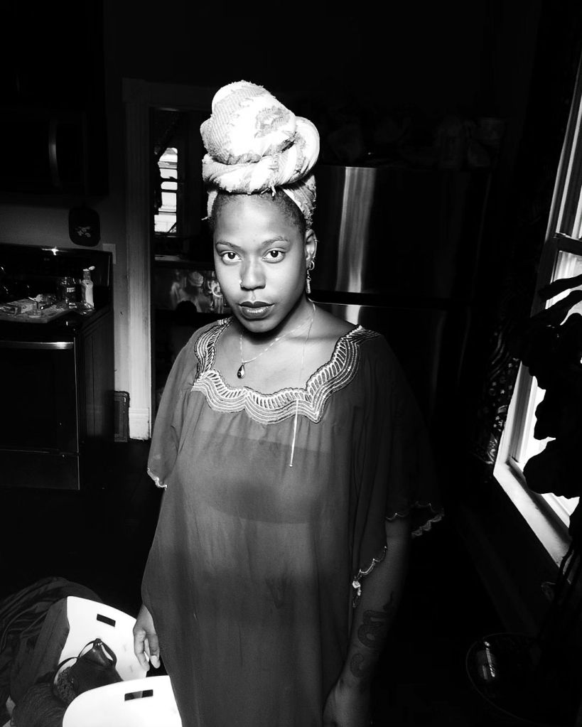 A Black woman stands in a dimly lit kitchen, gazing directly at the camera. She wears a sheer, flowing garment and a tall, intricately wrapped headwrap. The light from a nearby window softly illuminates her figure, casting gentle shadows behind her.