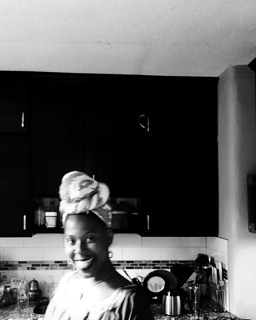 A smiling Black woman in a patterned headwrap stands in a softly lit kitchen, glowing with joy. The background is in deep shadow, highlighting her brightness.