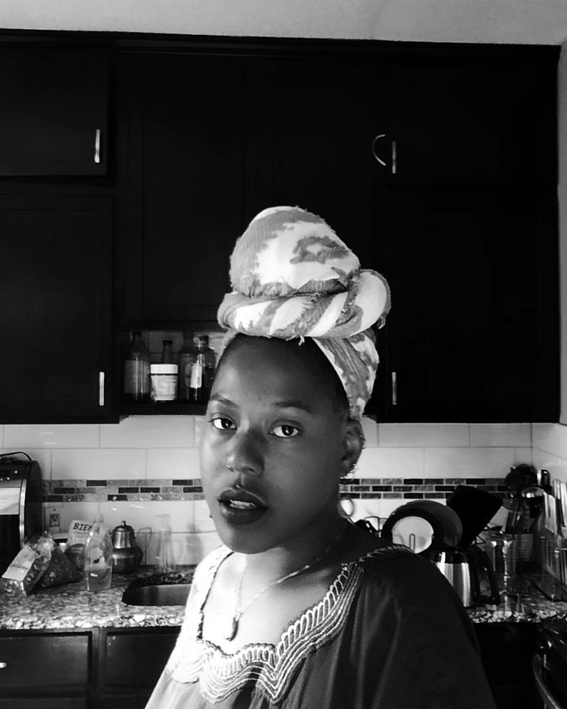 A Black woman in a kitchen, wrapped in a high headwrap and sheer traditional garment, gazes softly yet intensely toward the camera. The image is in black and white, creating contrast with the dark cabinets and reflecting kitchen surfaces behind her.