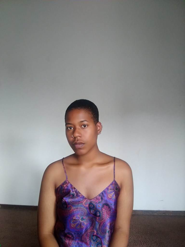 Black woman with a shaved head seated in front of a plain wall, wearing a purple paisley slip, gazing softly at the camera.