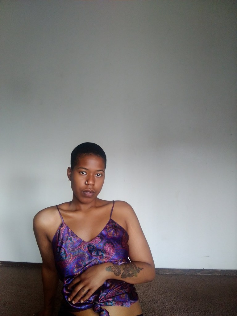 A woman with a shaved head sits in a purple patterned top, gazing softly into the camera against a plain wall, hand resting just above her belly.