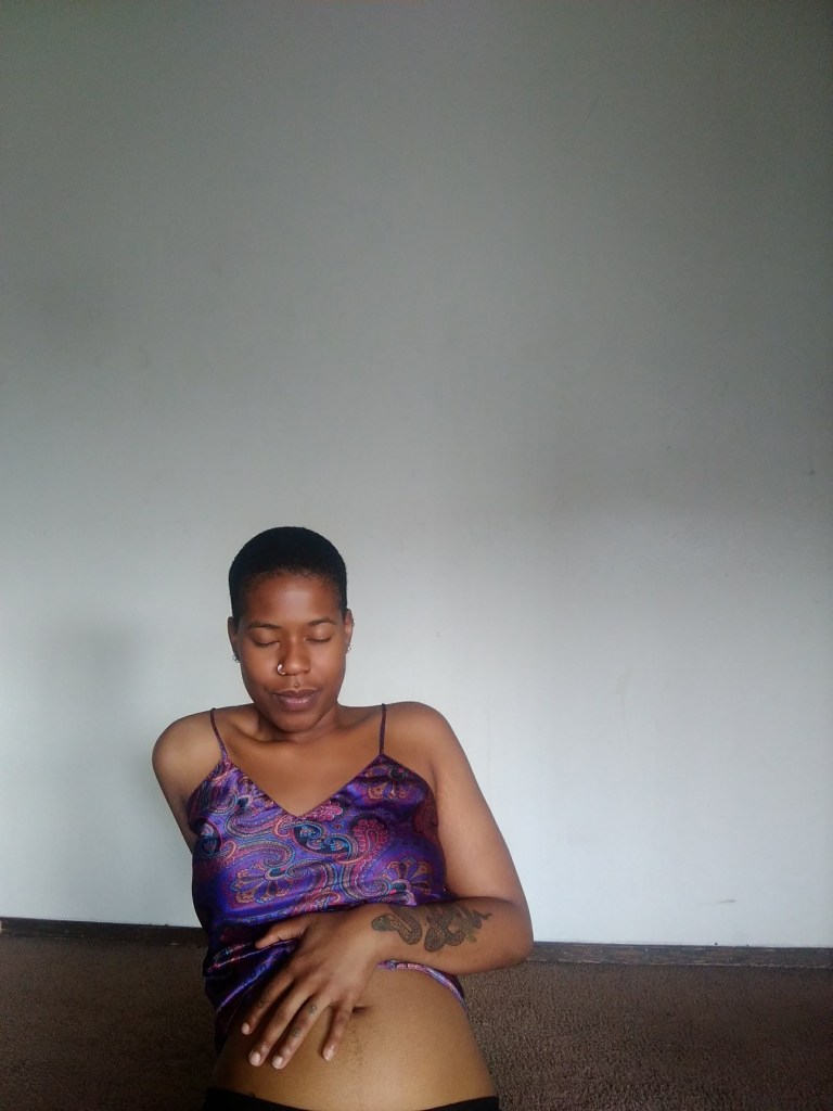 A woman in a purple satin top sits with eyes closed, one hand resting on her bare belly, against a blank white wall in soft natural light.