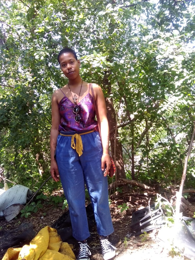 Woman standing in the woods, wearing blue pants, a purple top, and layered necklaces, surrounded by sunlight and overgrowth.