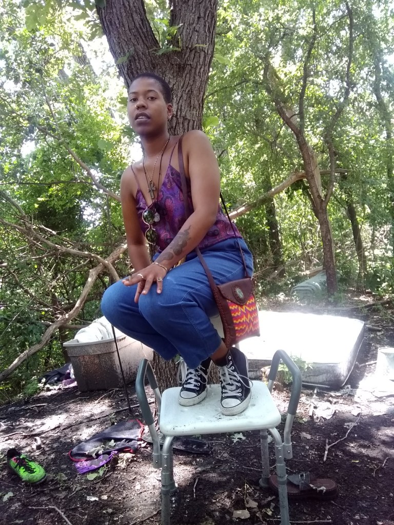 A woman balances on a metal chair in the forest, wearing a purple top, blue pants, and Converse sneakers.