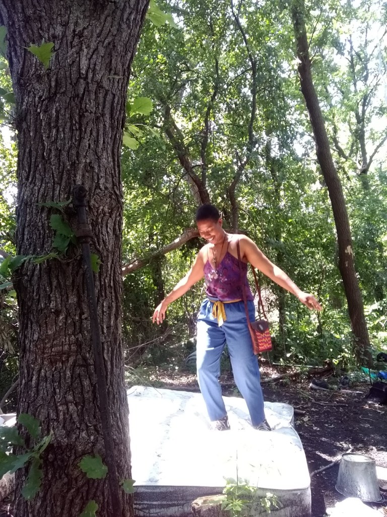 A woman stands with open arms on an old mattress in the woods, surrounded by sunlight and trees.