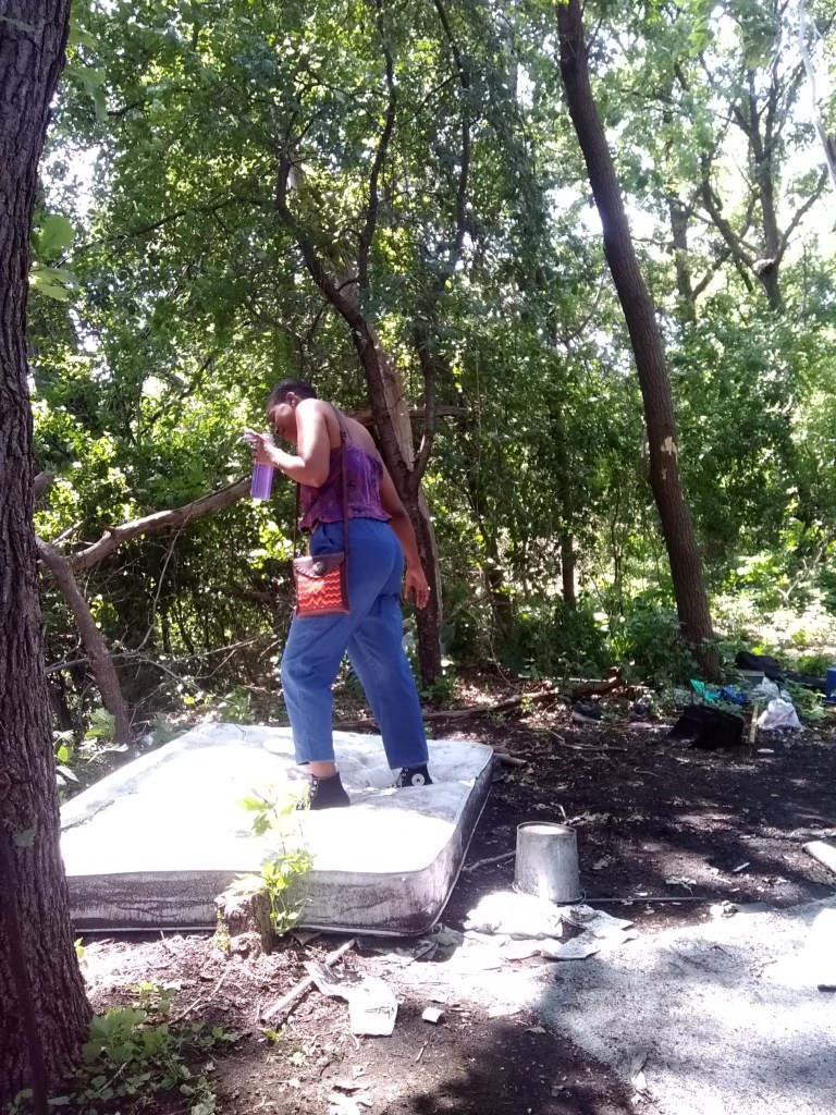 A woman stands on an old mattress in the woods, holding a drink, head bowed in motion beneath the trees.