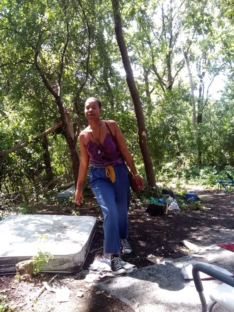 Woman smiling and leaning playfully in a sunlit forest, dressed in blue pants and a purple top.
