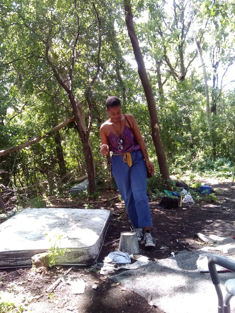 Woman in a purple top and blue pants standing on a stone in a wooded area, surrounded by nature and light.
