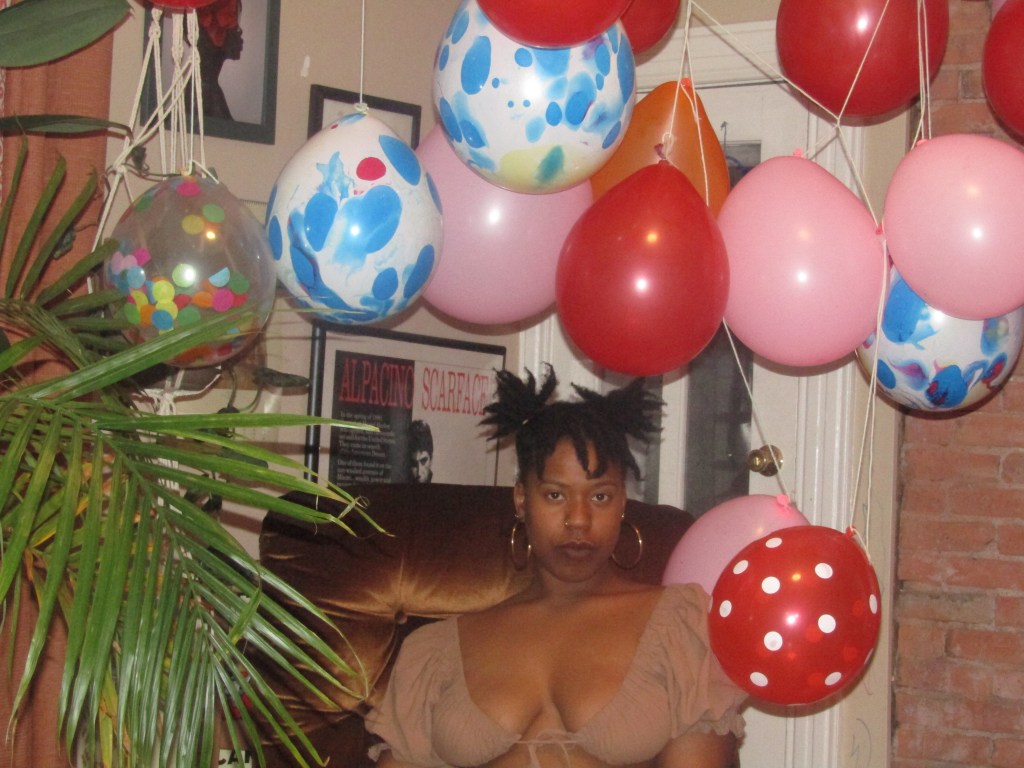 A woman sits calmly beneath a cascade of colorful balloons in a cozy, warmly lit room. She gazes directly at the camera with an air of quiet power and softness.