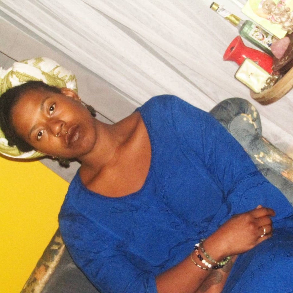 A Black woman sits slightly reclined on a patterned couch, wearing a rich cobalt blue dress and a light green-and-white headwrap. Her expression is grounded and contemplative. One arm rests across her body, bracelets and tattoos visible, against a mustard yellow wall and draped grey curtains. A shelf behind her holds candles, books.