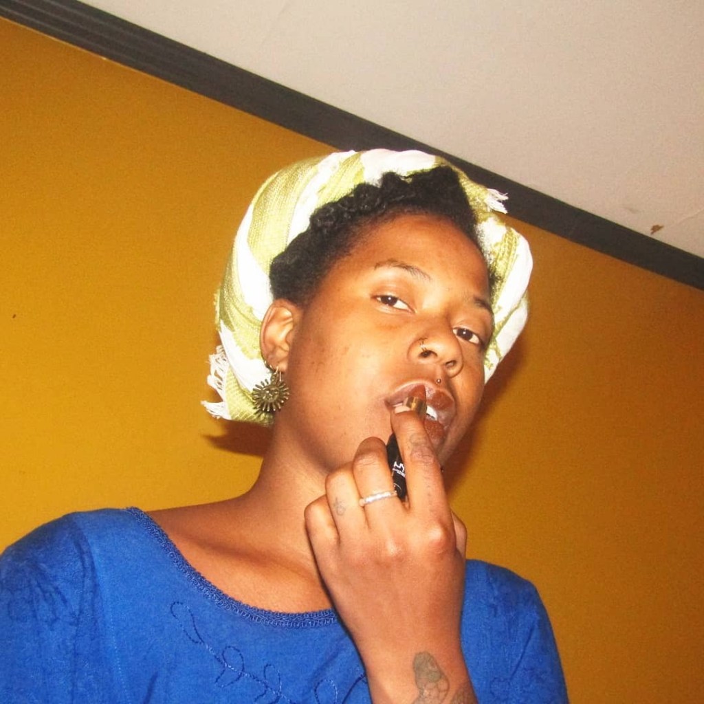 A Black woman in a vivid blue dress stands against a mustard yellow wall, gazing directly at the camera with a confident, serene expression. She wears a golden patterned headwrap and touches her lips with her fingers, mid-motion, evoking mystery and sensuality. A faint tattoo is visible on her forearm.