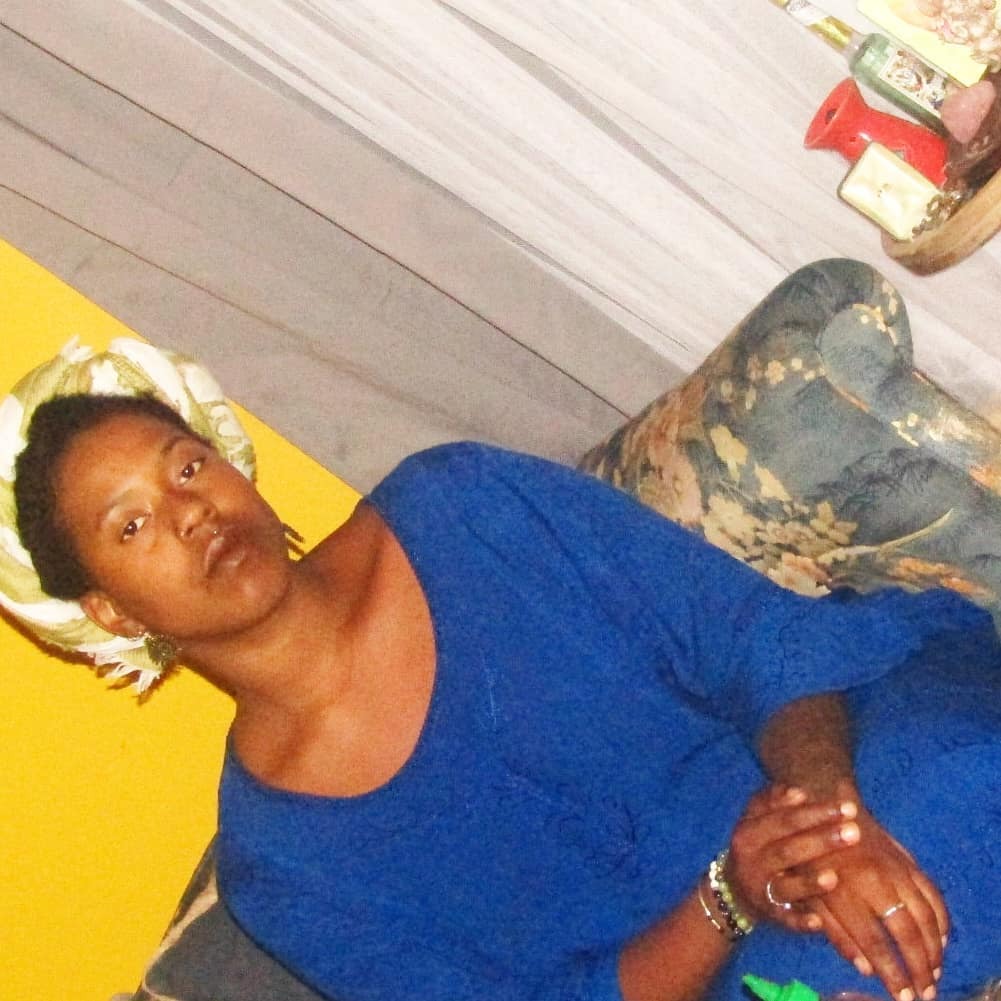 A Black woman sits on a patterned couch in a cozy, colorful room. She wears a deep blue dress and a light-colored headwrap, her hands gently clasped. Her expression is calm and composed, almost solemn. A mustard yellow wall and soft drapery frame the background, alongside books, candles, and lived-in textures.