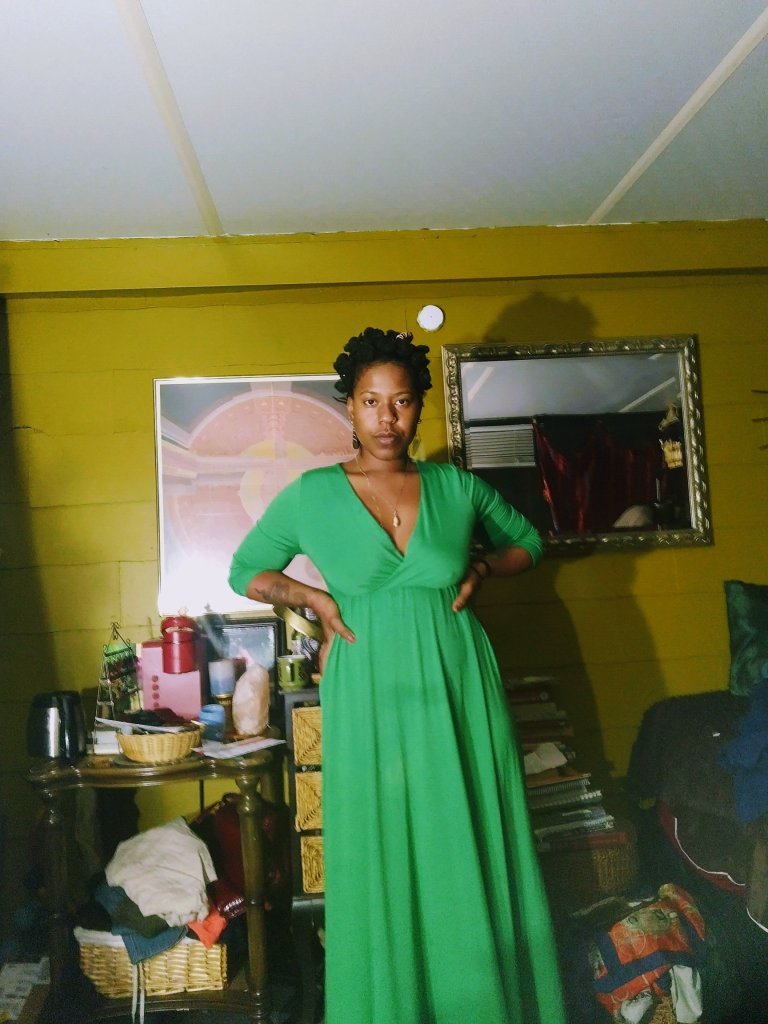 A woman in a flowing emerald green dress stands in a warmly lit room with golden walls, surrounded by books, baskets, and lived-in beauty. She gazes forward with quiet confidence, hands on hips.