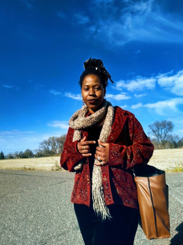 A woman stands on an open road in a red patterned jacket and scarf, carrying a tan bag. The sky above is vivid blue with scattered clouds, and a golden field stretches behind her.

