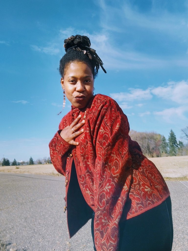 A woman in a red patterned jacket leans toward the camera with a soft, playful expression, standing on an open road beneath a blue sky.