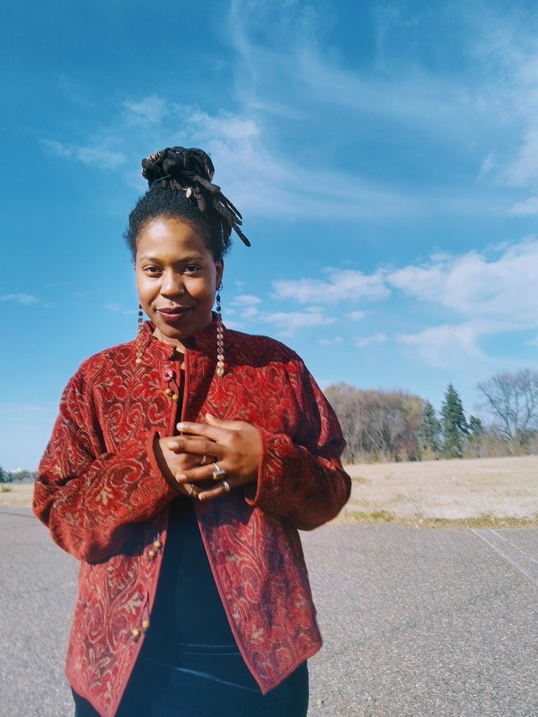 A woman stands under a vivid blue sky, wearing a red patterned jacket and looking into the camera with a gentle, knowing smile. Her hands are held near her heart.