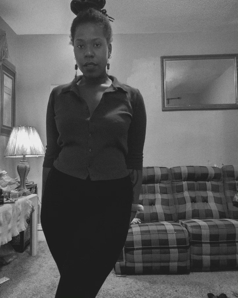 Black and white photo of a woman standing indoors with hands behind her back, facing forward. She wears a button-up top and long skirt, with a bun atop her head. A lamp glows softly to the side near a mirror and couch.