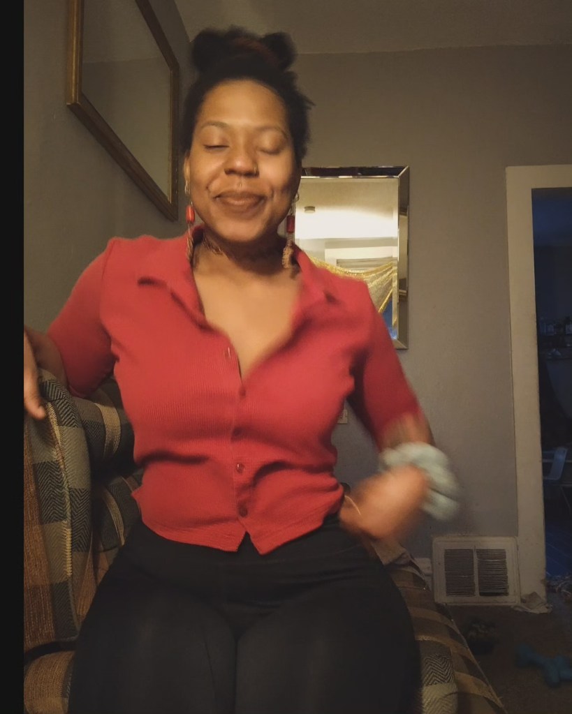 A woman sits on a patterned couch, smiling with her eyes closed, wearing a red blouse and black pants. The room is warmly lit with mirrors and a cozy, lived-in feel.