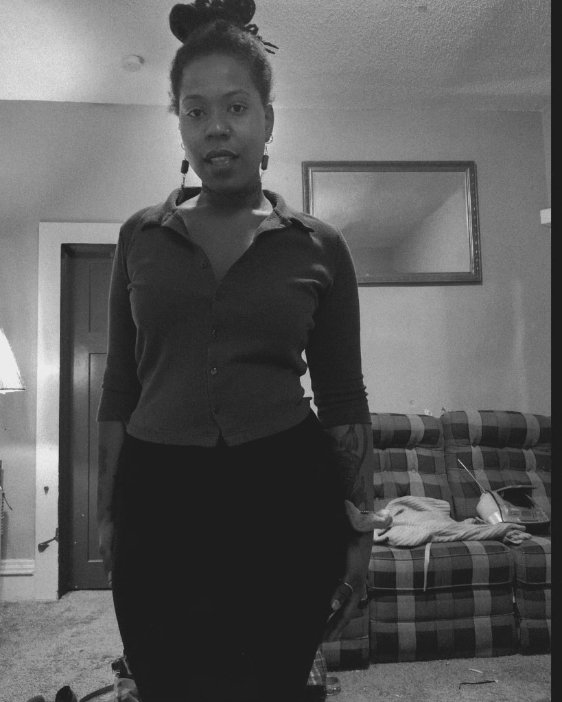 Black and white photo of a woman standing confidently indoors, wearing a button-up top with hair in a bun. A mirror and plaid couch are in the background.