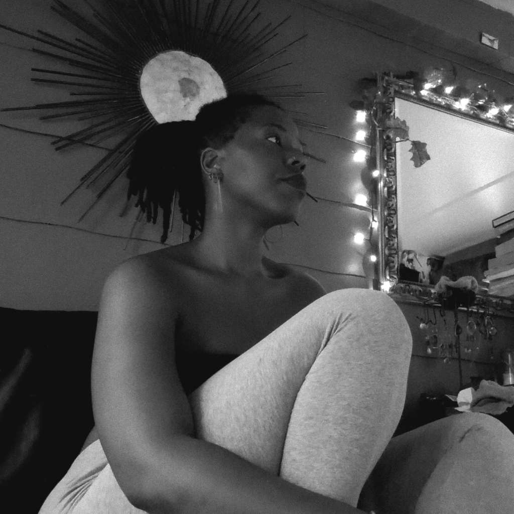 Black and white photo of a woman sitting beside a mirror, knee to chest, a radiant sunburst wall piece