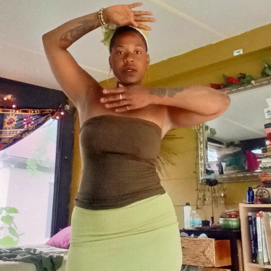 Woman standing indoors with one arm raised above her head and the other across her chest, in green clothing