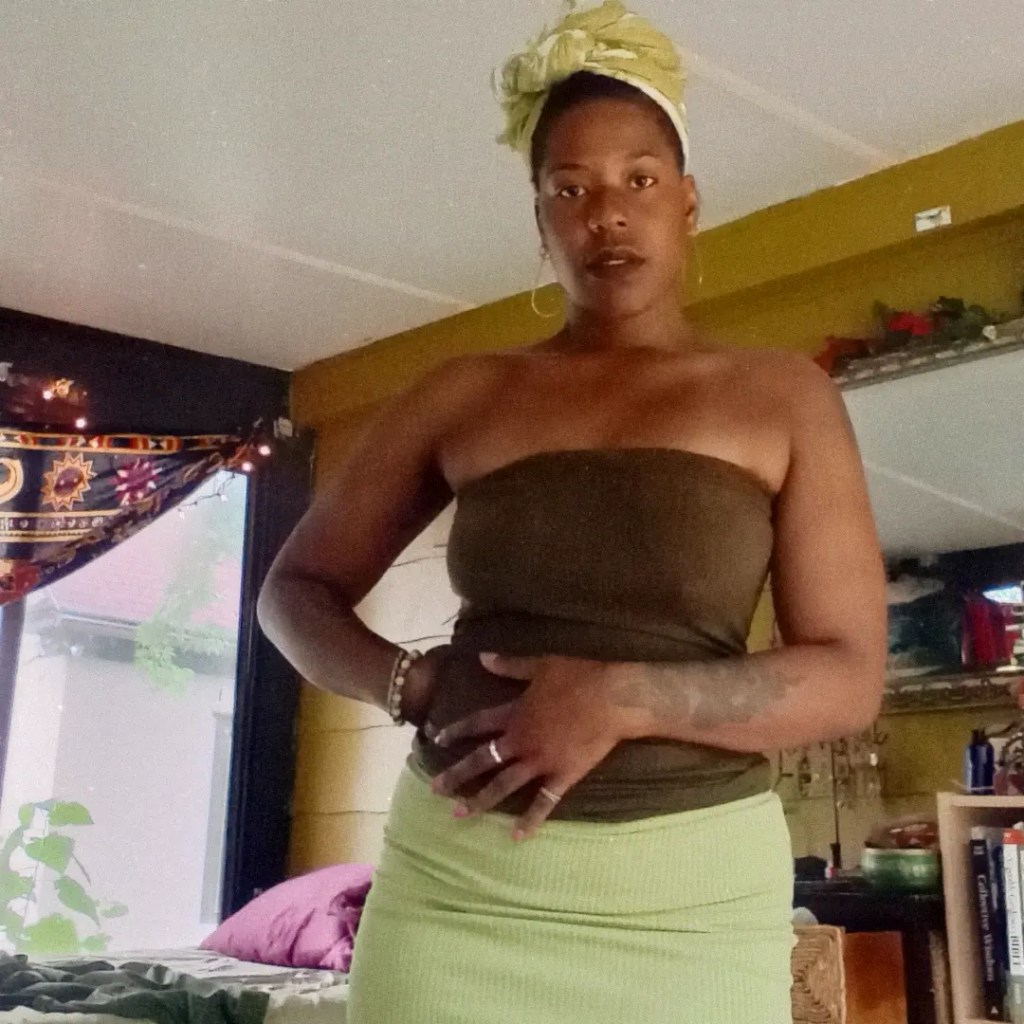 Woman standing indoors with both hands resting on her belly, gazing forward in a calm stance