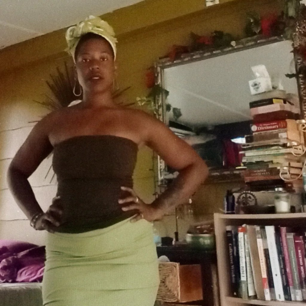 Woman indoors with hands on her hips, standing confidently near books and a mirror