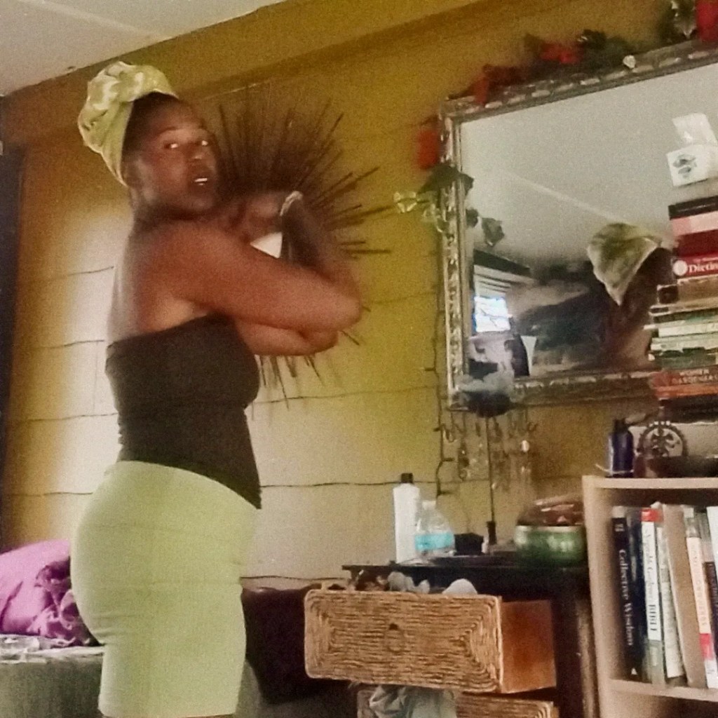 Woman turning slightly, indoors near a mirror and bookshelf, wearing a green headwrap.