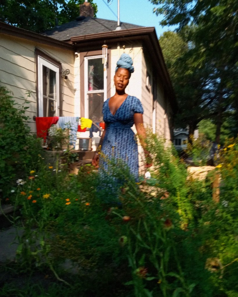 Woman in blue dress and headwrap standing in a lush, sunlit garden
