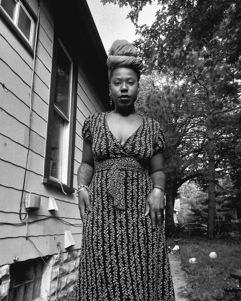 Black and white portrait of a woman standing tall in a patterned dress with a headwrap, outside house, surrounded by trees