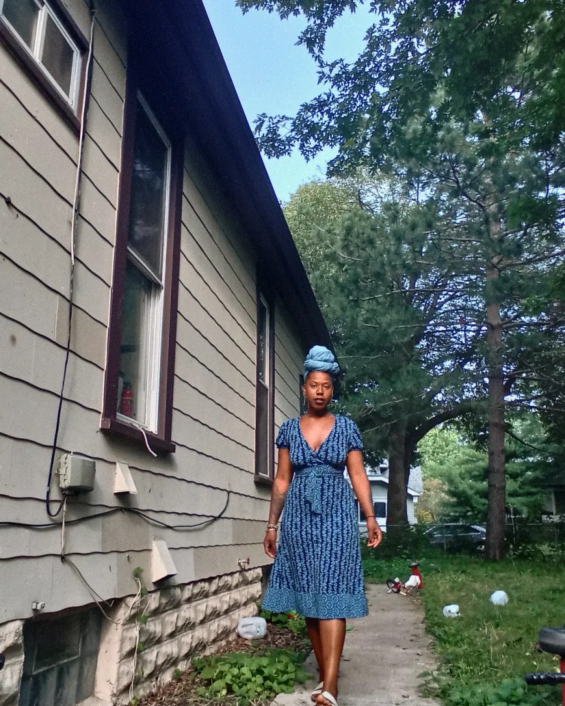 A woman in a blue dress and headwrap walks down a narrow sidewalk besides a house, with trees and soft daylight in the background