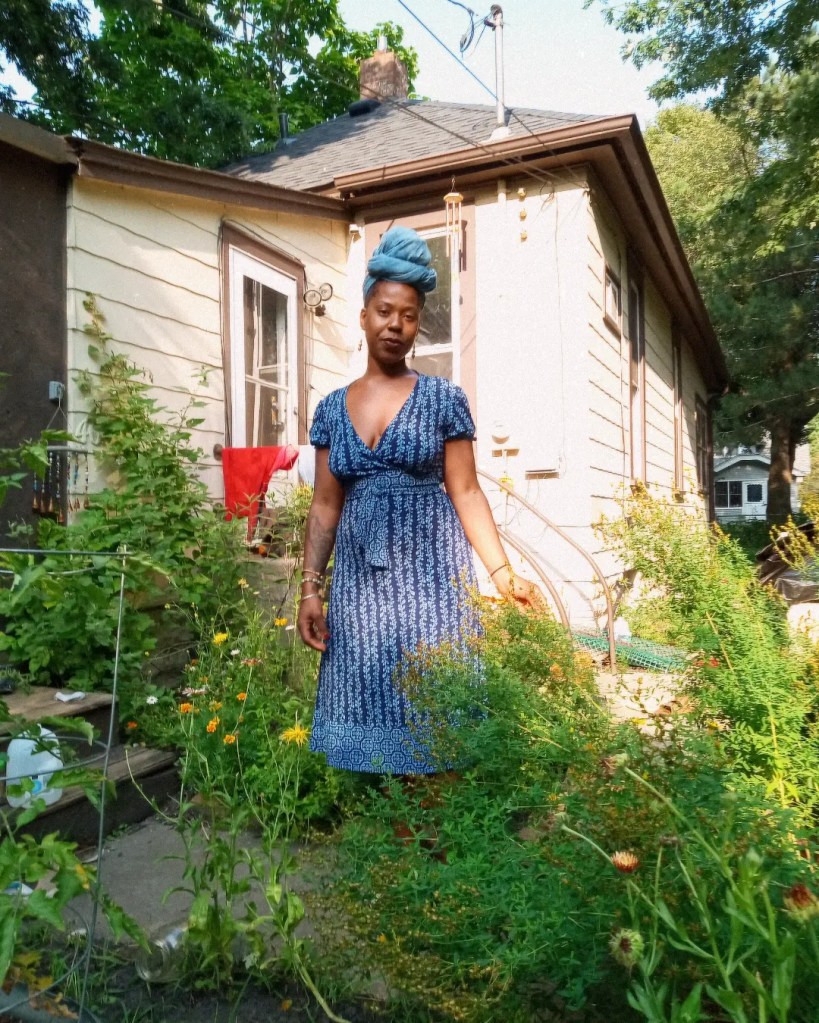 Woman in blue dress and headwrap standing in a lush garden, in front of a beige house, partially blurred in motion