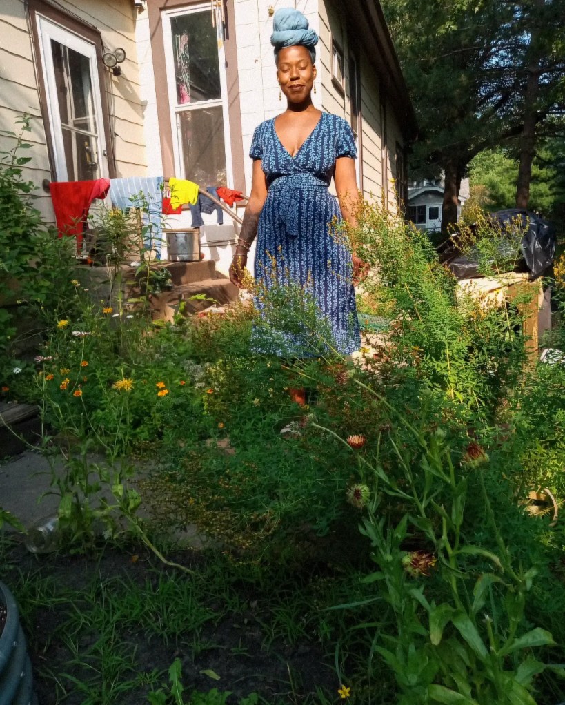 A woman in blue dress and headwrap stands among vibrant garden plants in front of a house, bathed in warm afternoon light