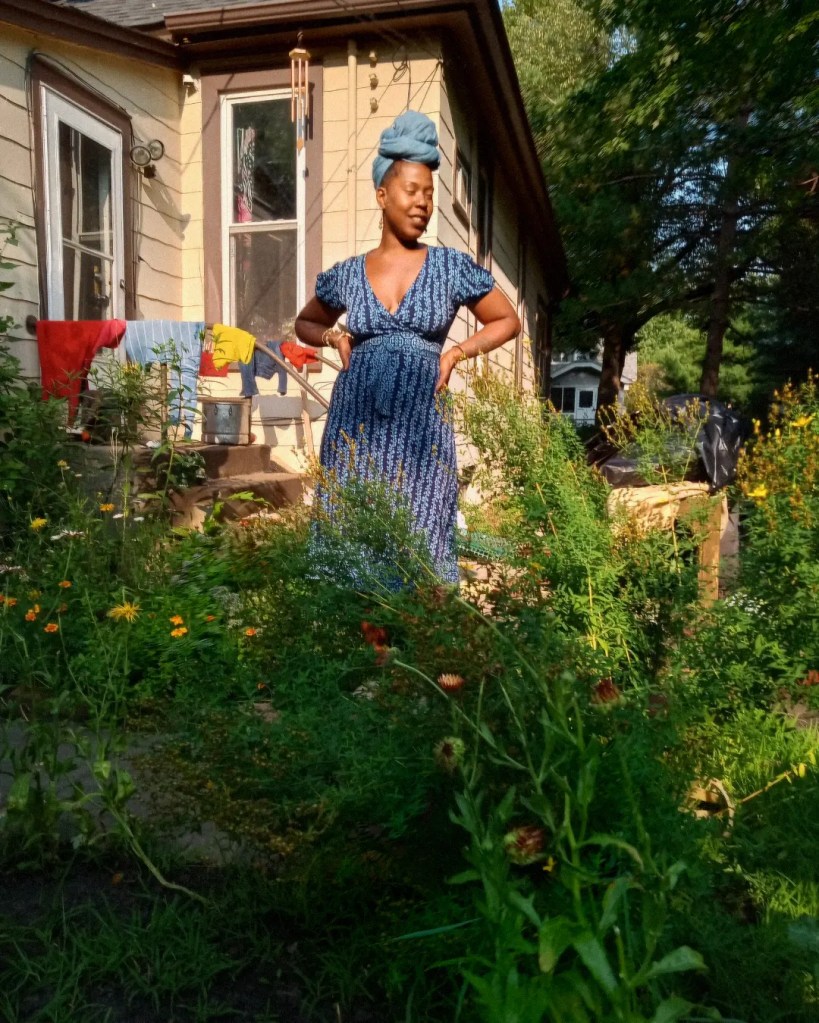 A woman in a blue dress and headwrap stands confidently among tall garden plants in front of a house, one hand on her hip, slightly blurred in motion