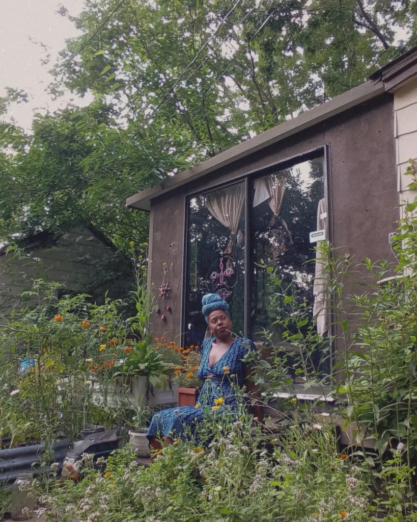 Seated woman in blue dress and headwrap, surrounded by garden plants in front of a house