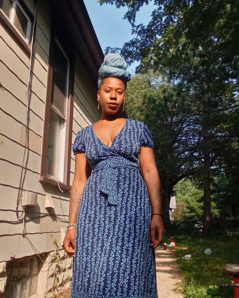 A woman in a blue patterned dress and headwrap stands outside house, softly lit by sunlight, with trees and yard in the background