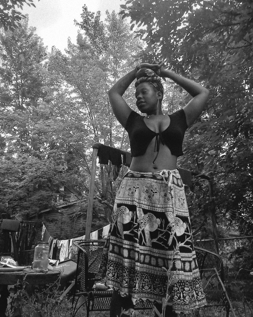 Black and white photo of a woman outdoors, standing with arms raised and crossed above her head, wearing a patterned skirt.