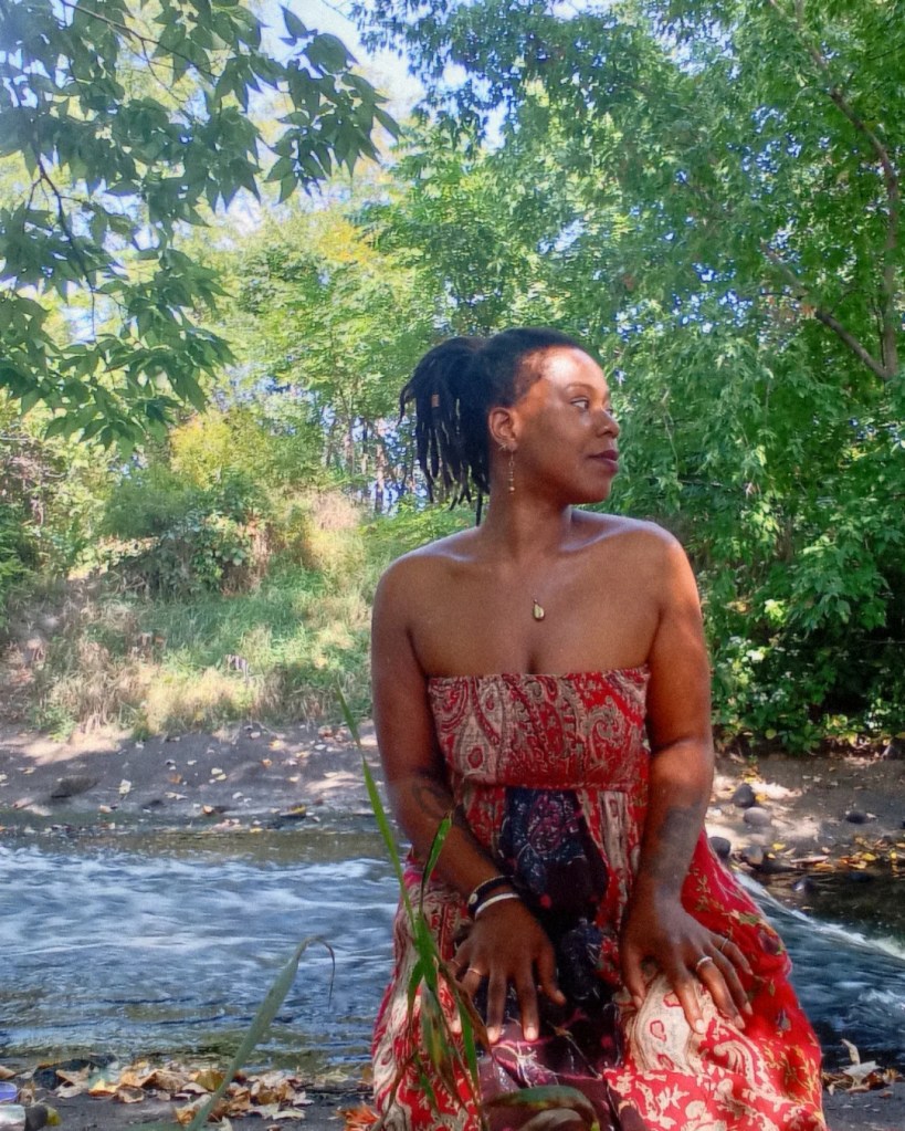 Woman in a red wrap dress sitting by the river, turned slightly, surrounded by greenery