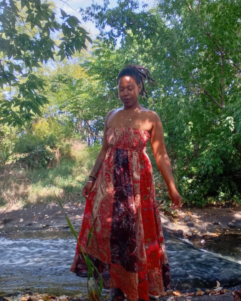 Woman in flowing red dress walking through a shallow stream, surrounded by lush greenery and sunlight
