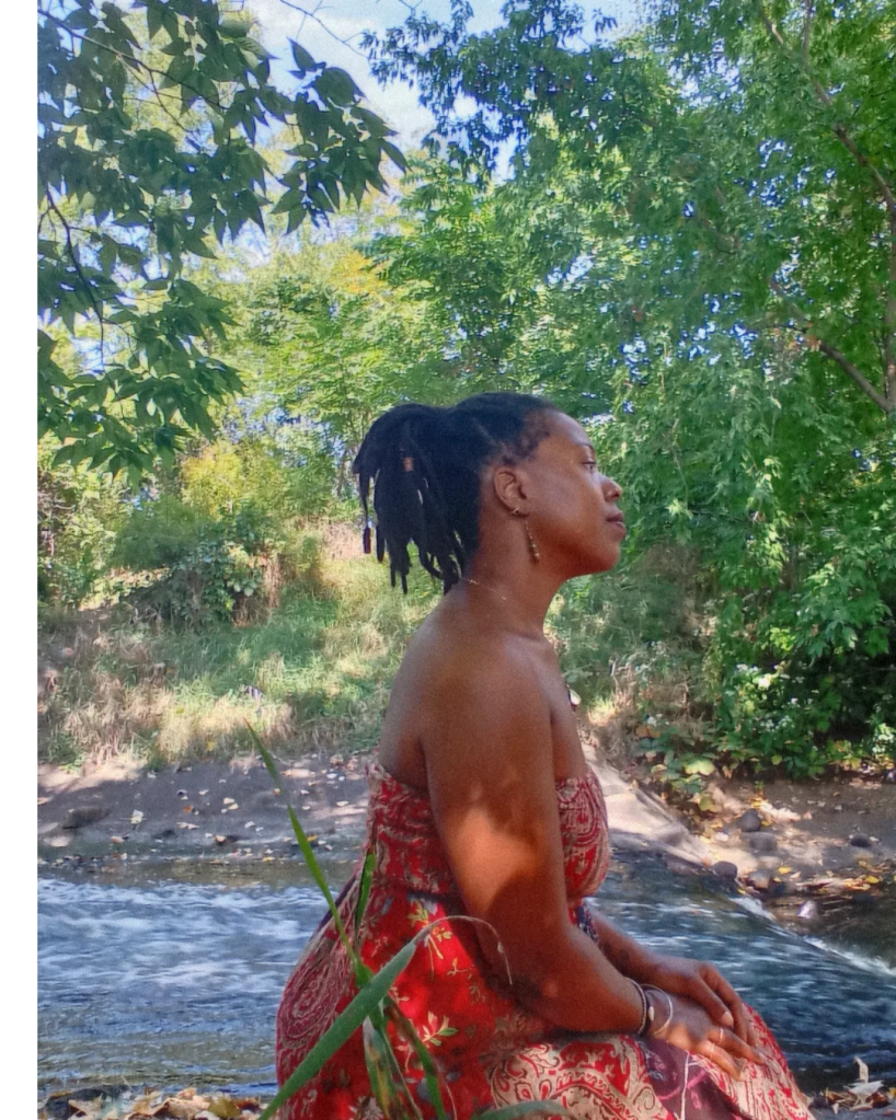 Woman in patterned dress sitting by a flowing river, in profile, surrounded by green trees