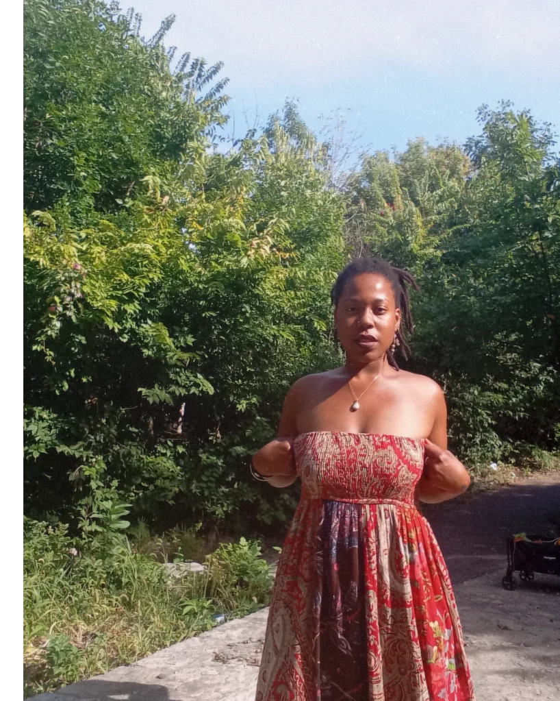 Woman in a patterned red dress standing in sunlight, adjusting her dress, surrounded by green foliage