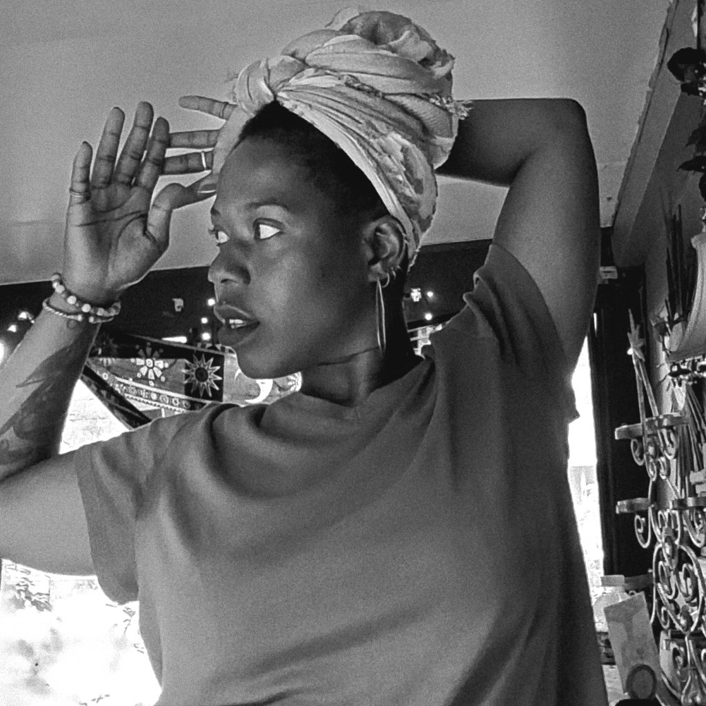 Black and white photo of woman indoors, head wrapped, gazing off to the side with one arm lifted.