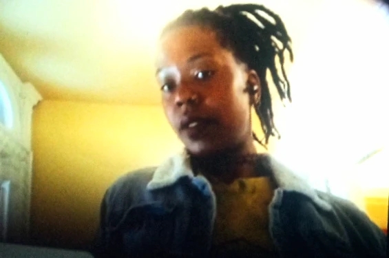 Soft-focus portrait of a woman with locs, wearing a denim jacket, against a golden wall
